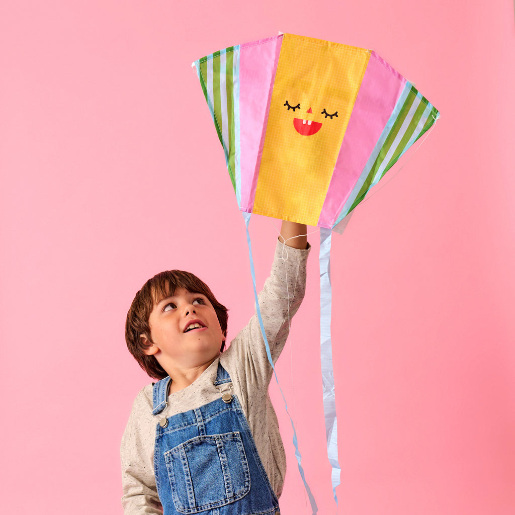 A young child in denim overalls joyfully holds a colorful kite with a smiling face design against a bright pink background. The scene is playful and cheerful.