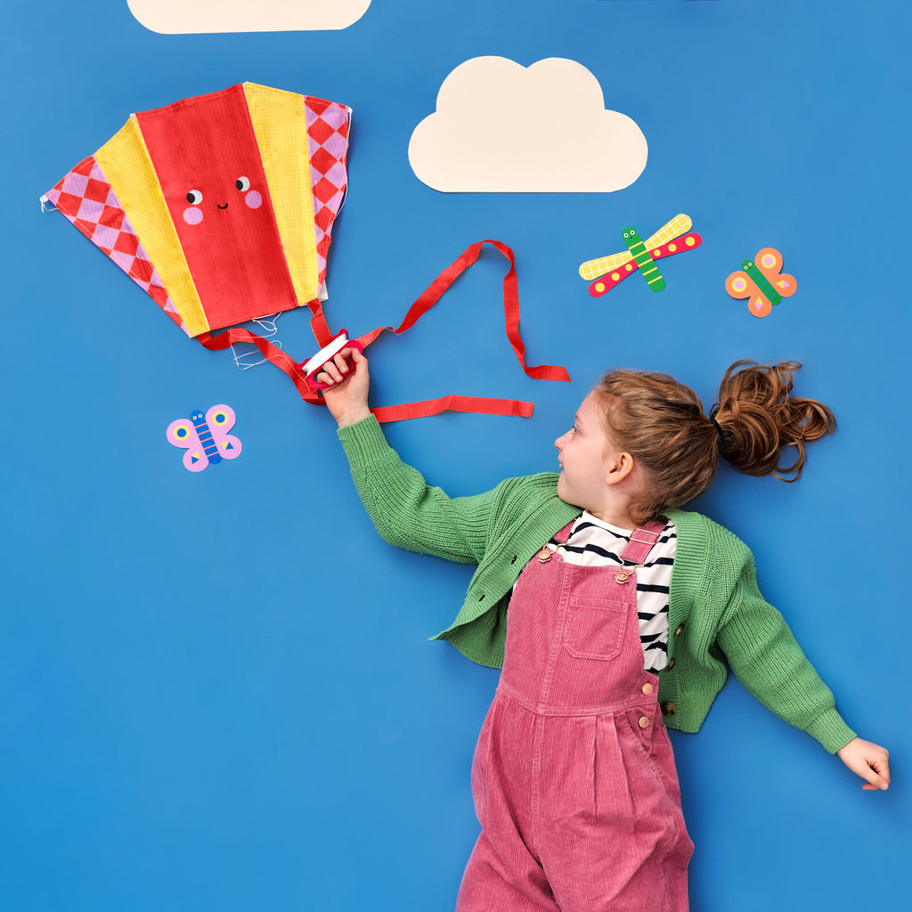 A girl in pink overalls and a green sweater holds a colorful kite against a blue backdrop with white clouds and paper butterflies, appearing joyful.