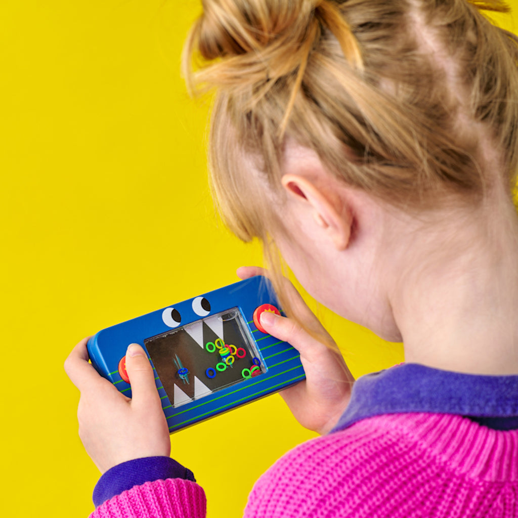 A young girl in a pink sweater plays a handheld ring toss game against a bright yellow background, focusing intently on the colorful rings.