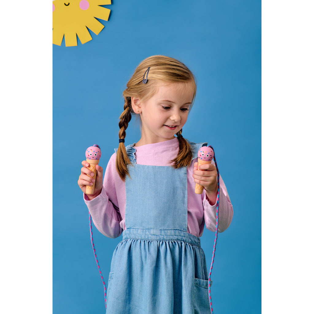 A young girl in denim overalls and a pink shirt holds two pink ice cream jump rope handles against a blue background with a smiling sun decoration.