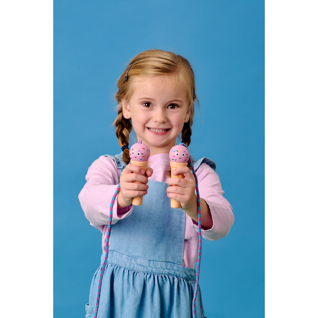 A smiling child in a denim dress and pink shirt holds two ice cream-shaped toys with colorful sprinkles, set against a blue background.