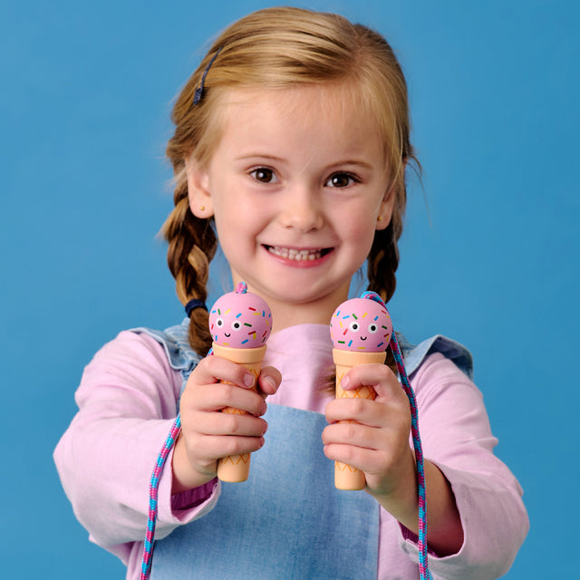 A smiling child in a denim dress and pink shirt holds two ice cream-shaped toys with colorful sprinkles, set against a blue background.