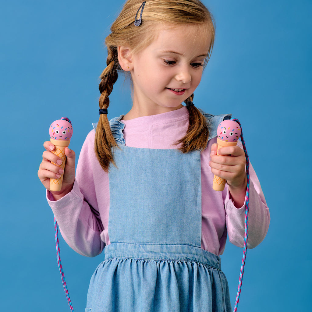 A young girl in denim overalls and a pink shirt holds two pink ice cream jump rope handles against a blue background with a smiling sun decoration.