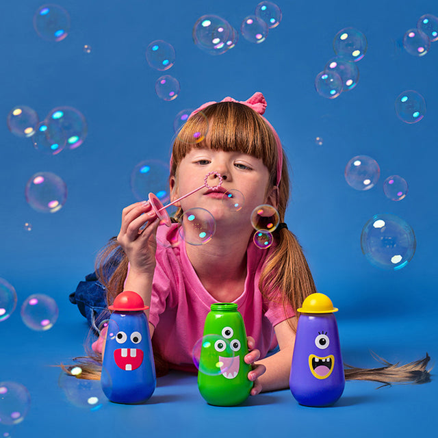 A girl in a pink shirt and headband joyfully blows bubbles. Colorful bottles with cartoon faces surround her against a bright blue background.