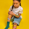 A child with braided hair sits against a yellow backdrop, wearing a white shirt and rolled-up jeans. Colorful band-aids cover their knees, adding a playful tone.