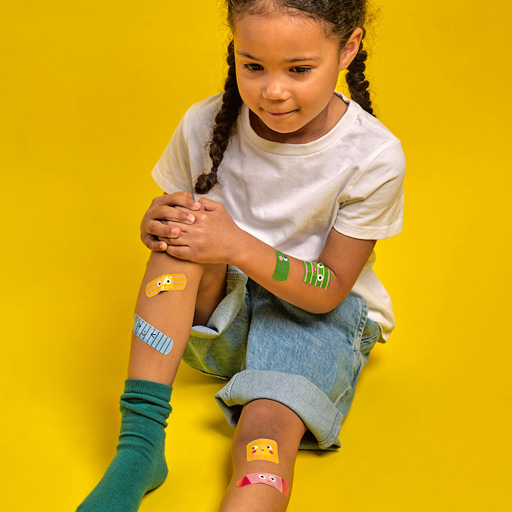 A child with braided hair sits against a yellow backdrop, wearing a white shirt and rolled-up jeans. Colorful band-aids cover their knees, adding a playful tone.