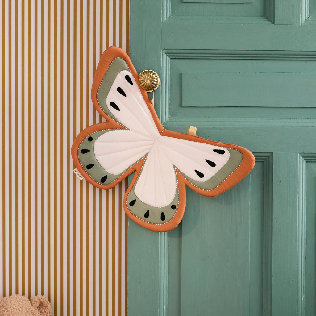 Fabric butterfly wings costume in soft orange, cream, and sage green with black teardrop accents. Two yellow straps suggest they can be worn. Bright and whimsical design.