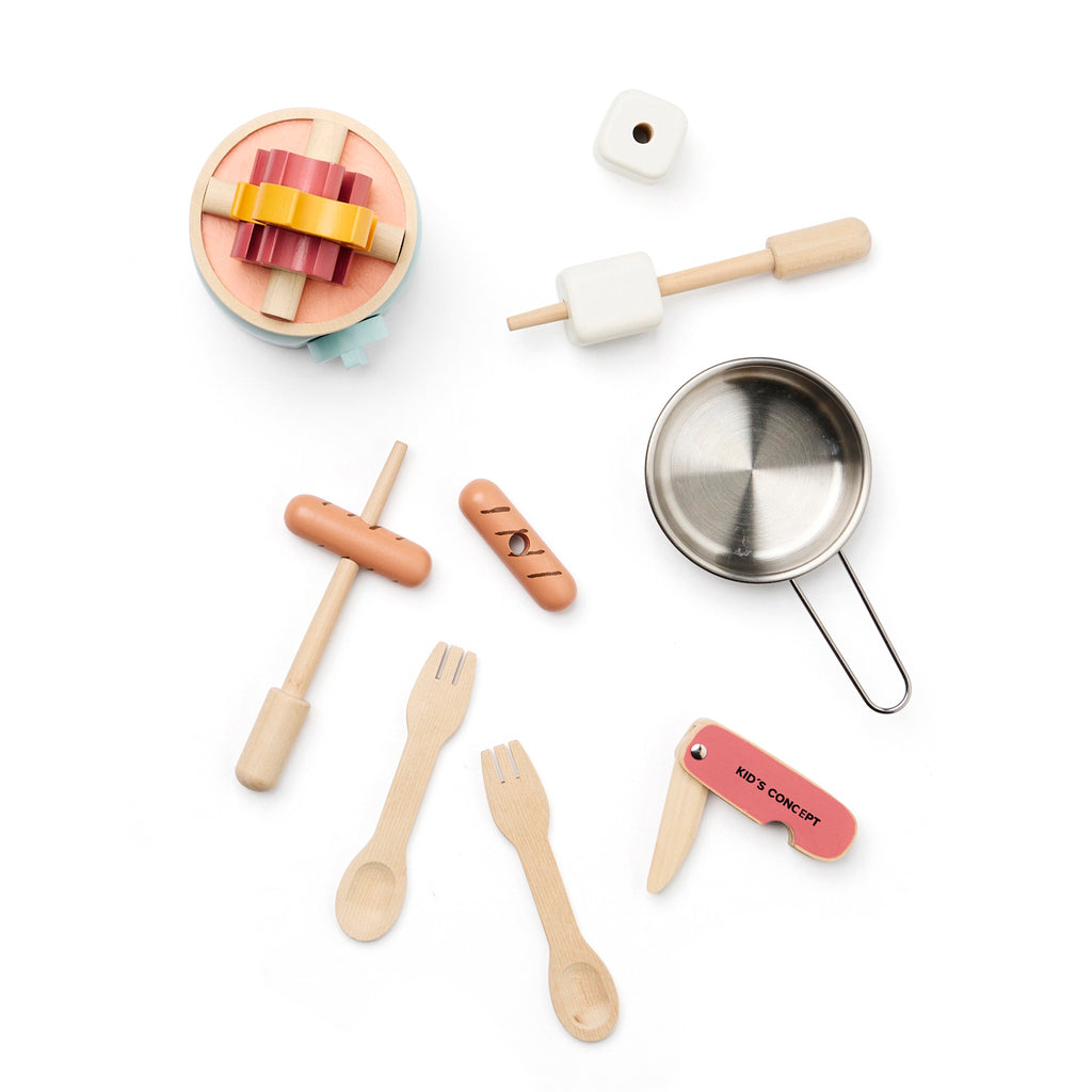 Wooden toy kitchen utensils include a pot on a mint stove, cutlery, a pretend food set, and a toy campfire, all arranged playfully, in white background.