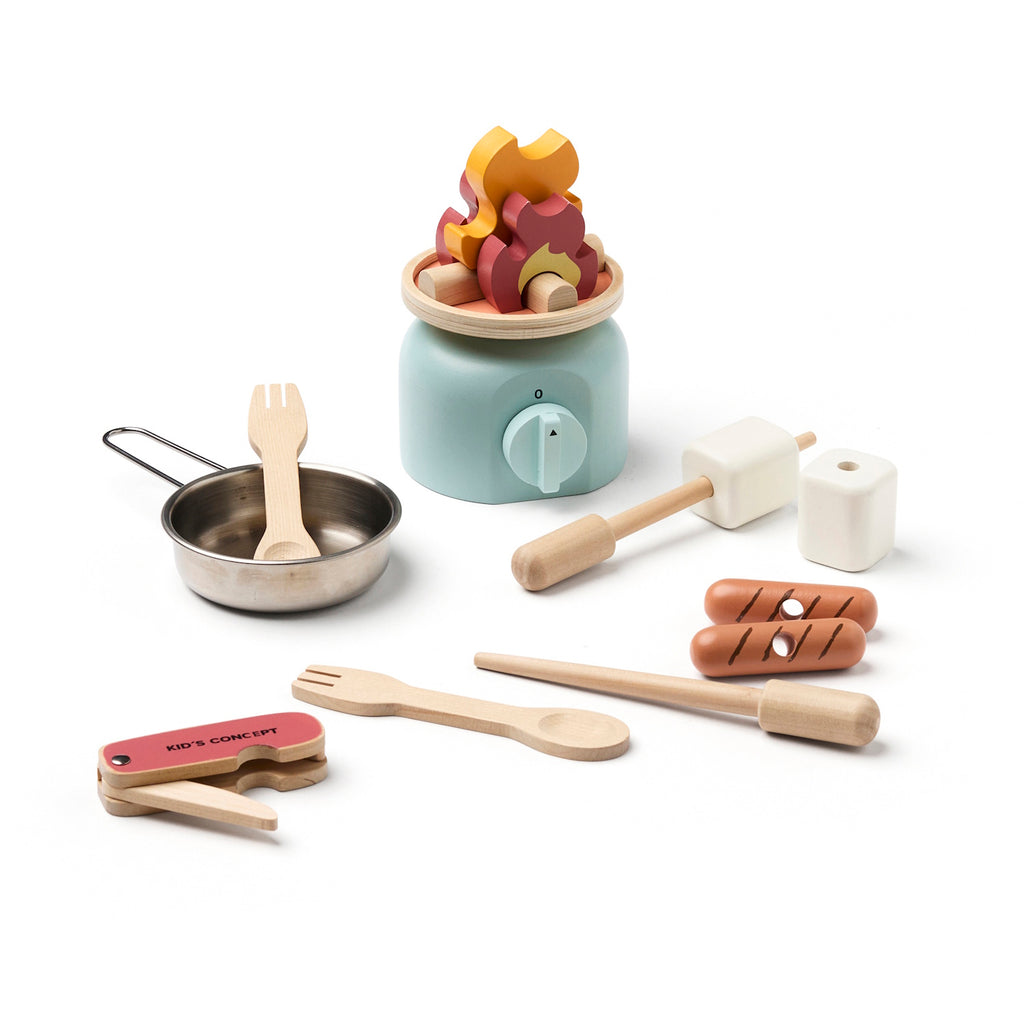 Wooden toy kitchen utensils include a pot on a mint stove, cutlery, a pretend food set, and a toy campfire, all arranged playfully, in white background.