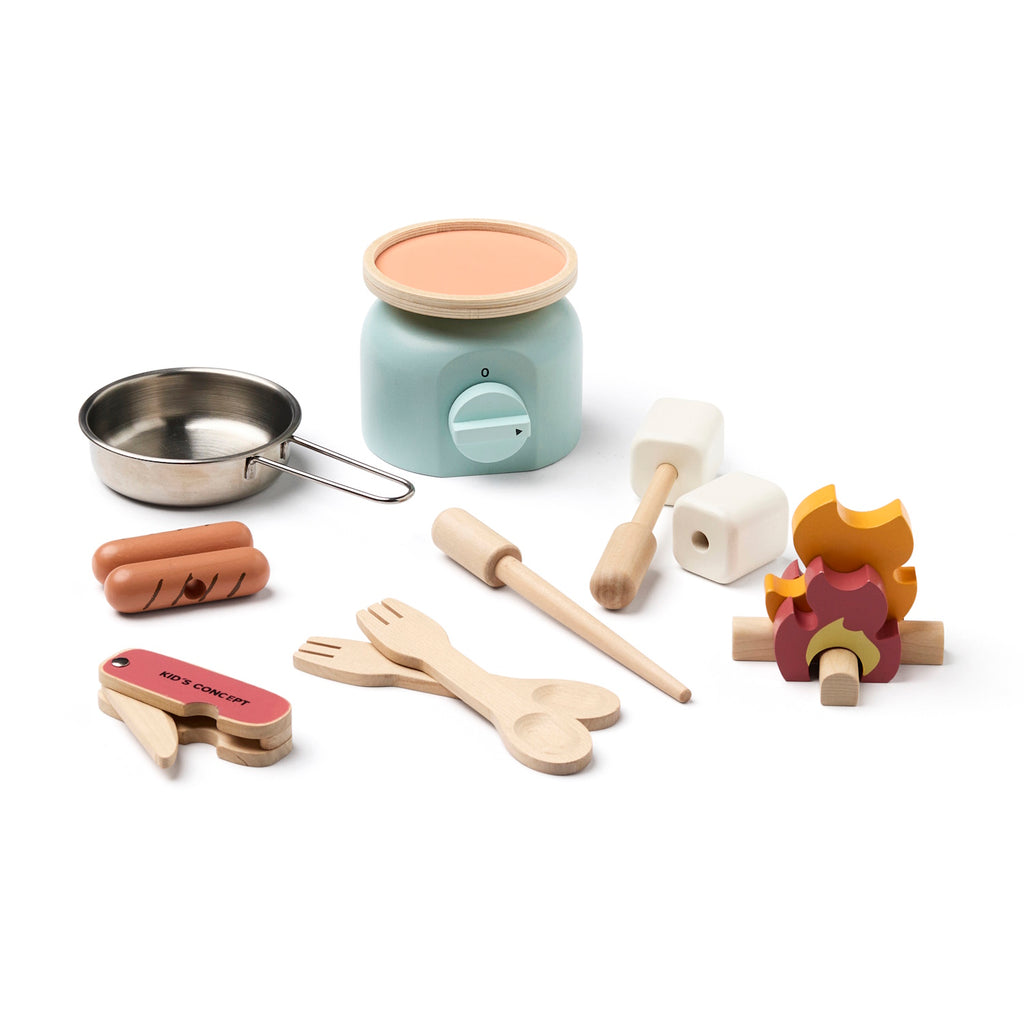 Wooden toy kitchen utensils include a pot on a mint stove, cutlery, a pretend food set, and a toy campfire, all arranged playfully, in white background.