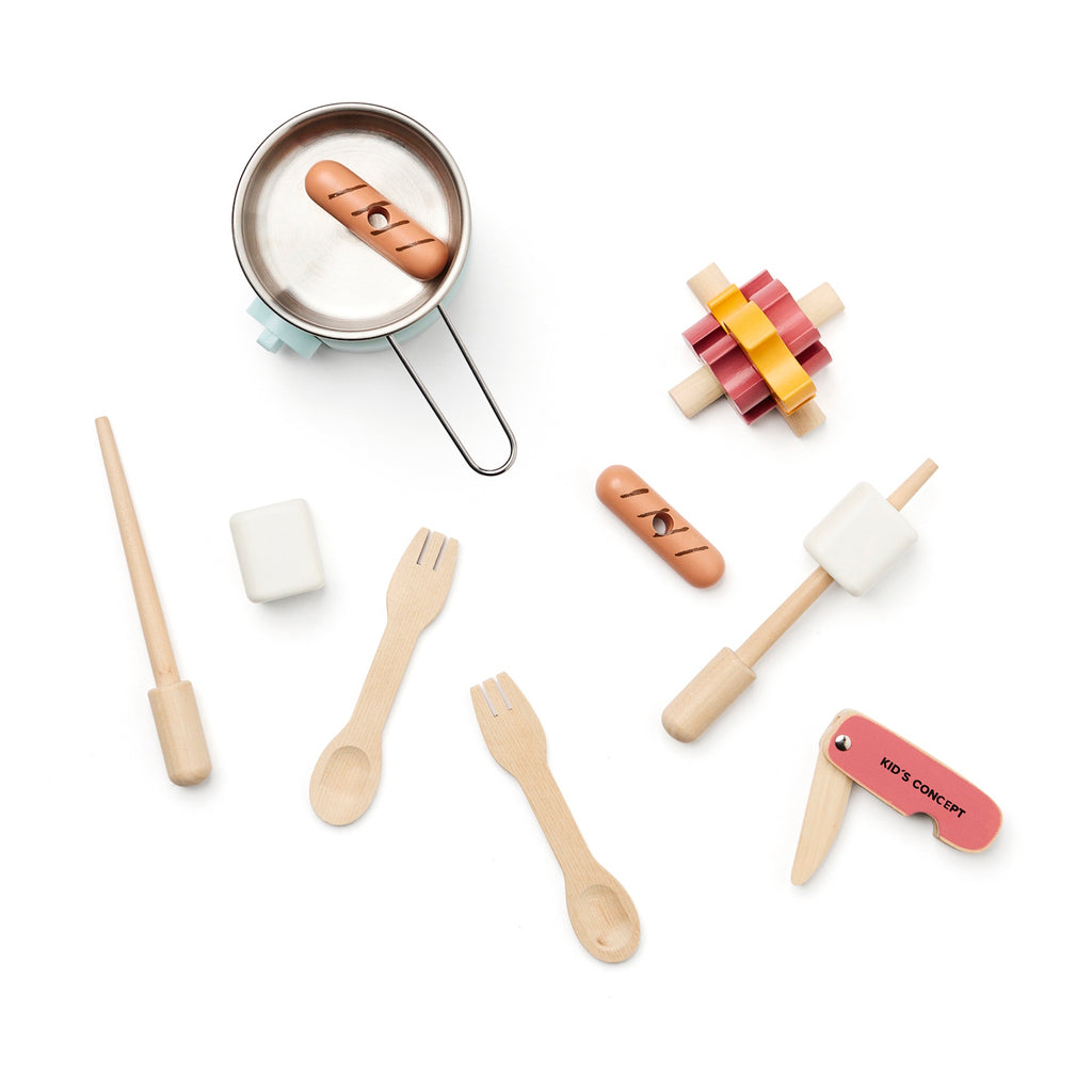 Wooden toy kitchen utensils include a pot on a mint stove, cutlery, a pretend food set, and a toy campfire, all arranged playfully, in white background.