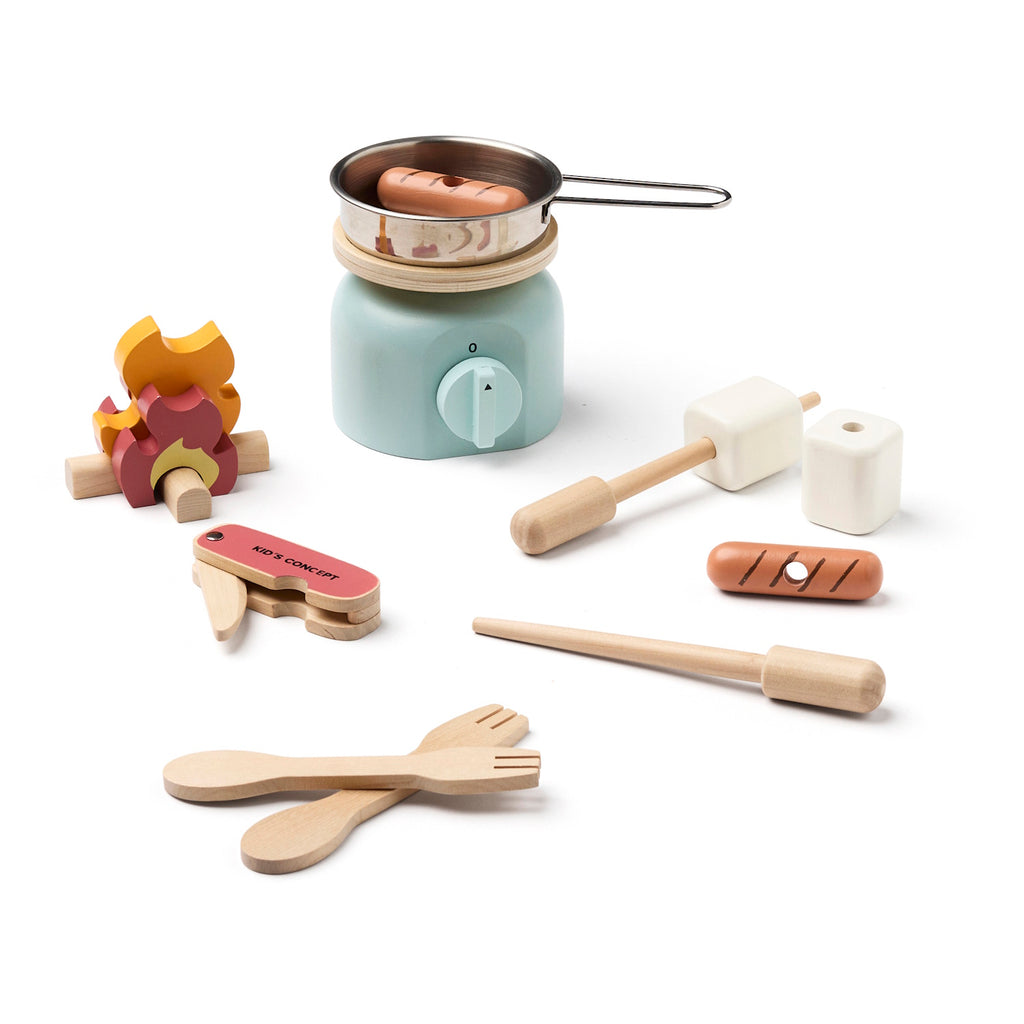 Wooden toy kitchen utensils include a pot on a mint stove, cutlery, a pretend food set, and a toy campfire, all arranged playfully, in white background.