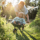 An adult giving a barefoot toddler in a sweater a ride inside a green wheelbarrow through a grassy garden outdoors.