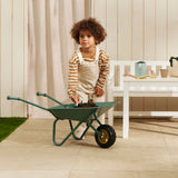 Child using a hand shovel to dig in a green wheelbarrow filled with soil near a white bench.