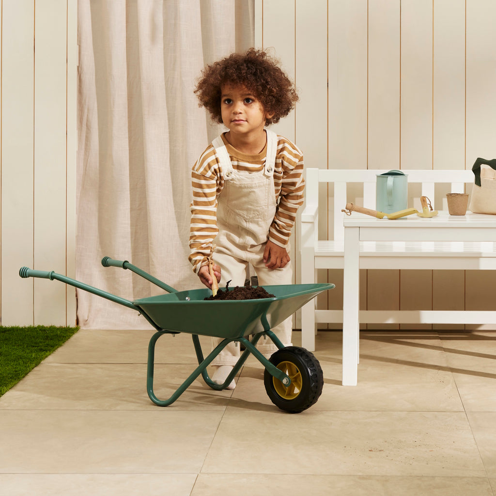 A green metal wheelbarrow with a single black rubber wheel and two handles, for gardening and construction pretend play.