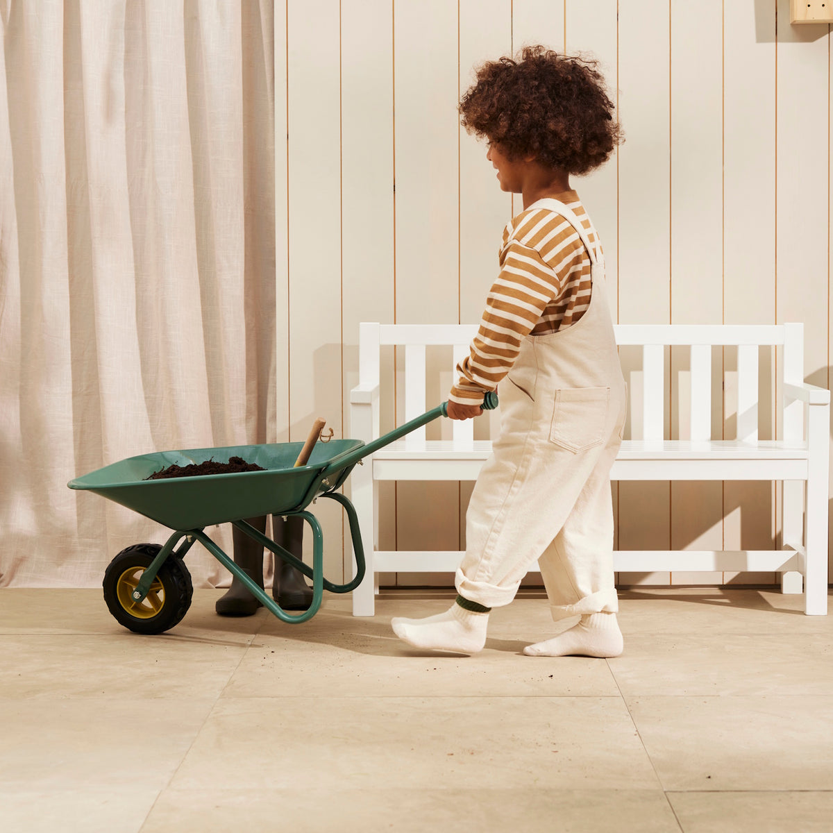 Child pushing a green wheelbarrow filled with soil past a white wooden bench.