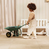 Child pushing a green wheelbarrow filled with soil past a white wooden bench.