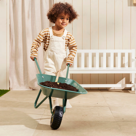 Young child pushing a green wheelbarrow filled with soil directly towards the camera.