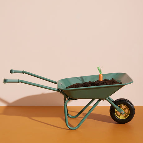 A green wheelbarrow filled with soil, with a single toy wooden carrot sticking out from the top, against a plain background.