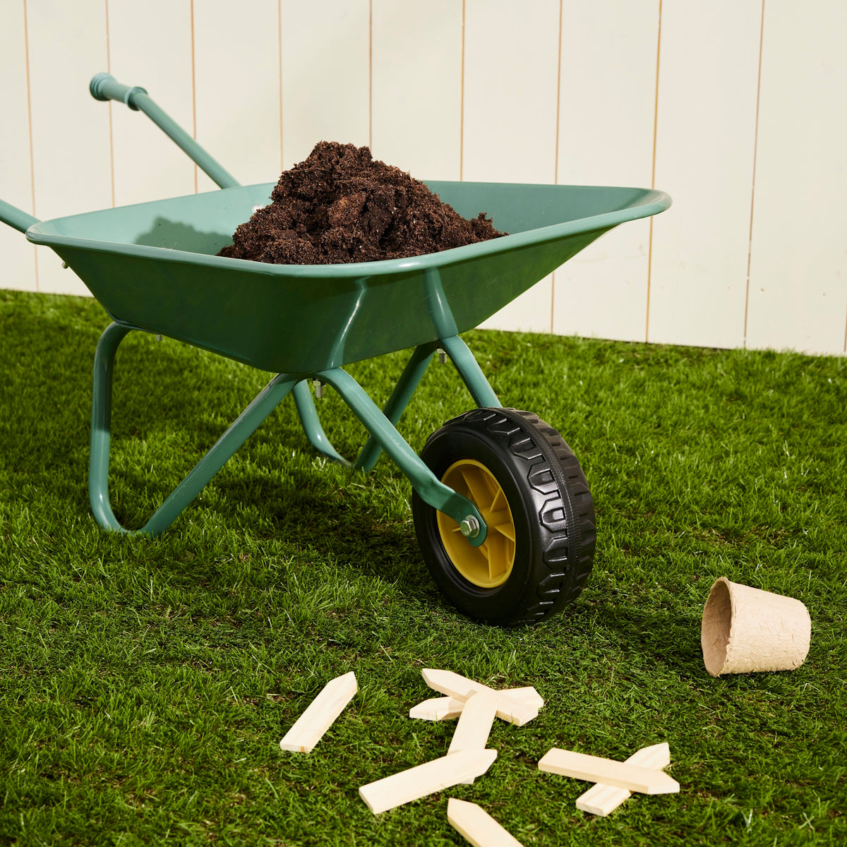 Green toy wheelbarrow filled with soil on grass with wooden plant markers nearby.