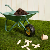 Green toy wheelbarrow filled with soil on grass with wooden plant markers nearby.