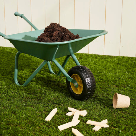 Green toy wheelbarrow filled with soil on grass with wooden plant markers nearby.