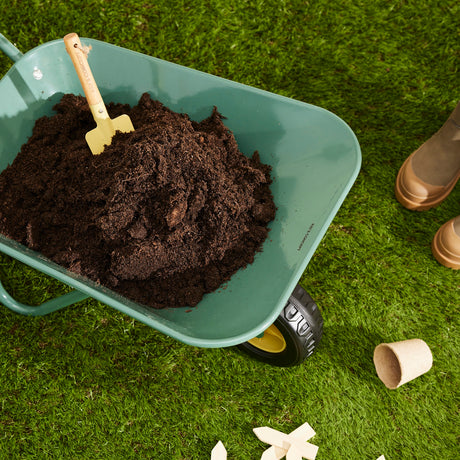 Green toy wheelbarrow filled with soil and a small trowel resting on green grass.