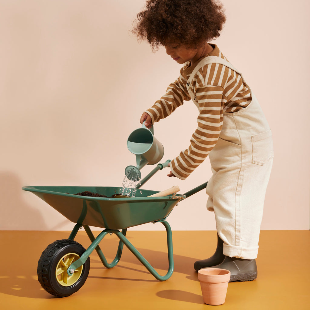 A green metal wheelbarrow with a single black rubber wheel and two handles, for gardening and construction pretend play.