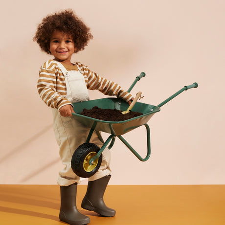 Smiling child wearing overalls and boots holding up a green wheelbarrow filled with soil.