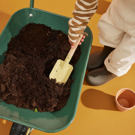 High-angle shot of a child scooping soil out of a green wheelbarrow using a small yellow hand shovel.