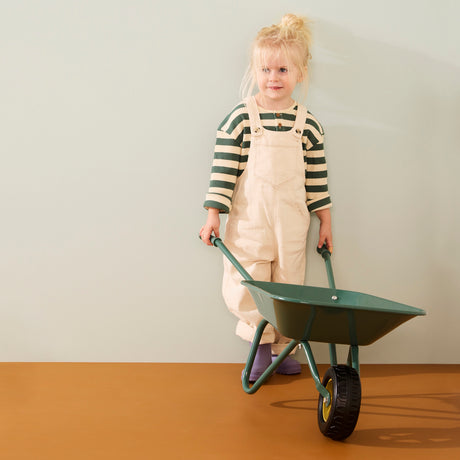 Young blonde child wearing cream overalls and a green striped shirt holding the handles of a green wheelbarrow.