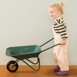 Young blonde child wearing cream overalls, a green striped shirt, and purple boots pushing a green wheelbarrow.