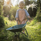 Young blonde toddler wearing striped overalls pushing a green wheelbarrow through a grassy garden filled with flowers.