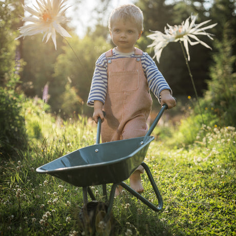 Young blonde toddler wearing striped overalls pushing a green wheelbarrow through a grassy garden filled with flowers.