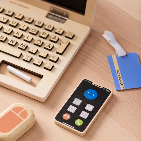 Wooden toy laptop play set with a toy smartphone, mouse, and ID badge on a desk.