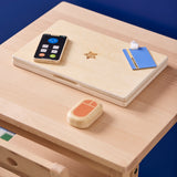 Closed wooden toy laptop, smartphone, mouse, and ID badge set resting on a wooden desk.