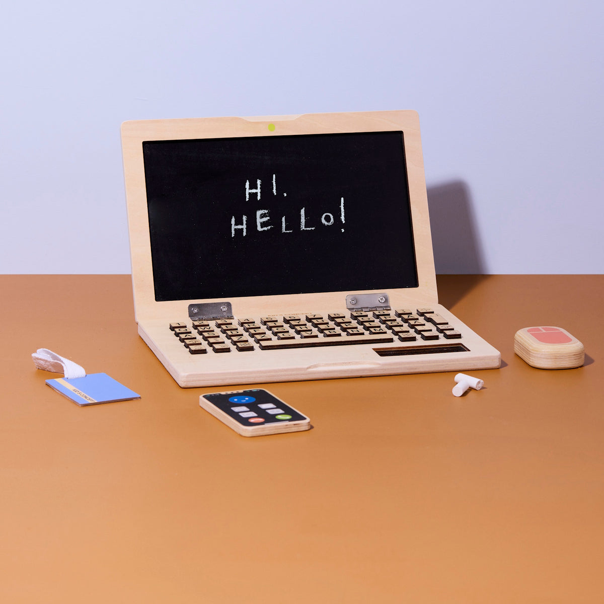 Wooden toy laptop with chalk writing on screen, wooden phone, mouse, chalk, and card on a desk.