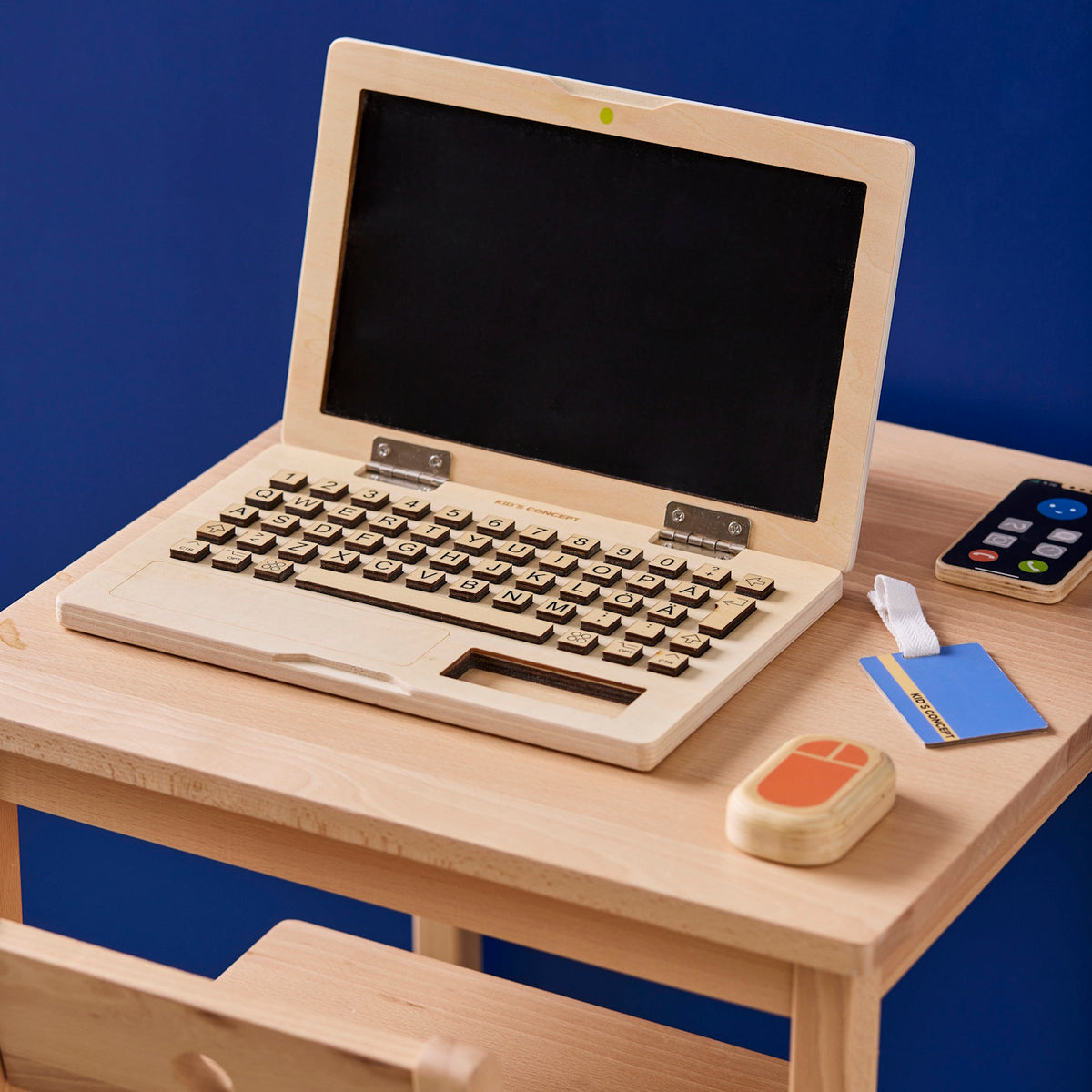 A wooden toy laptop play set on a desk with a wooden mouse, phone, and ID badge.