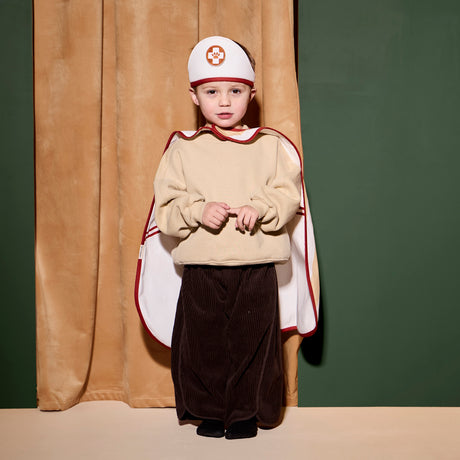 A child wearing a doctor dress-up headband and cape standing in front of curtains.