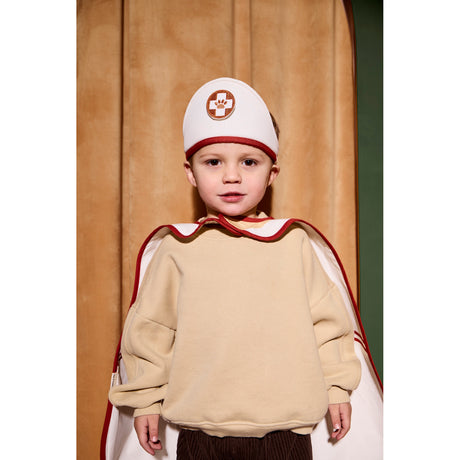 Child wearing a doctor dress-up hat and red-trimmed cape in front of a curtain.