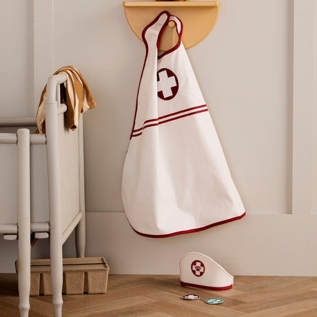 White doctor cape with red cross hanging on wall hook, with cap and badges on floor below.