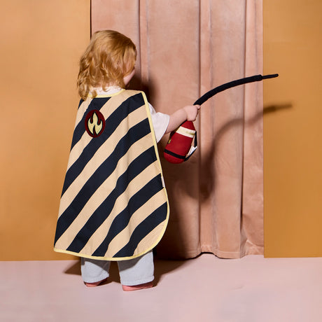 Child from behind in a striped cape with a flame emblem, holding a soft toy fire extinguisher.