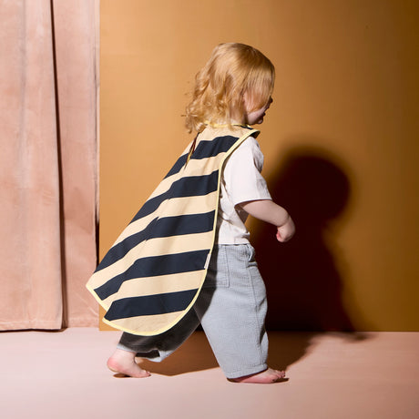 Toddler with blonde curly hair walking in profile while wearing a striped dress-up cape and gray trousers.