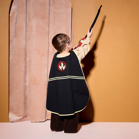 Child from behind wearing a black firefighter cape with a flame logo, holding up a toy hose.