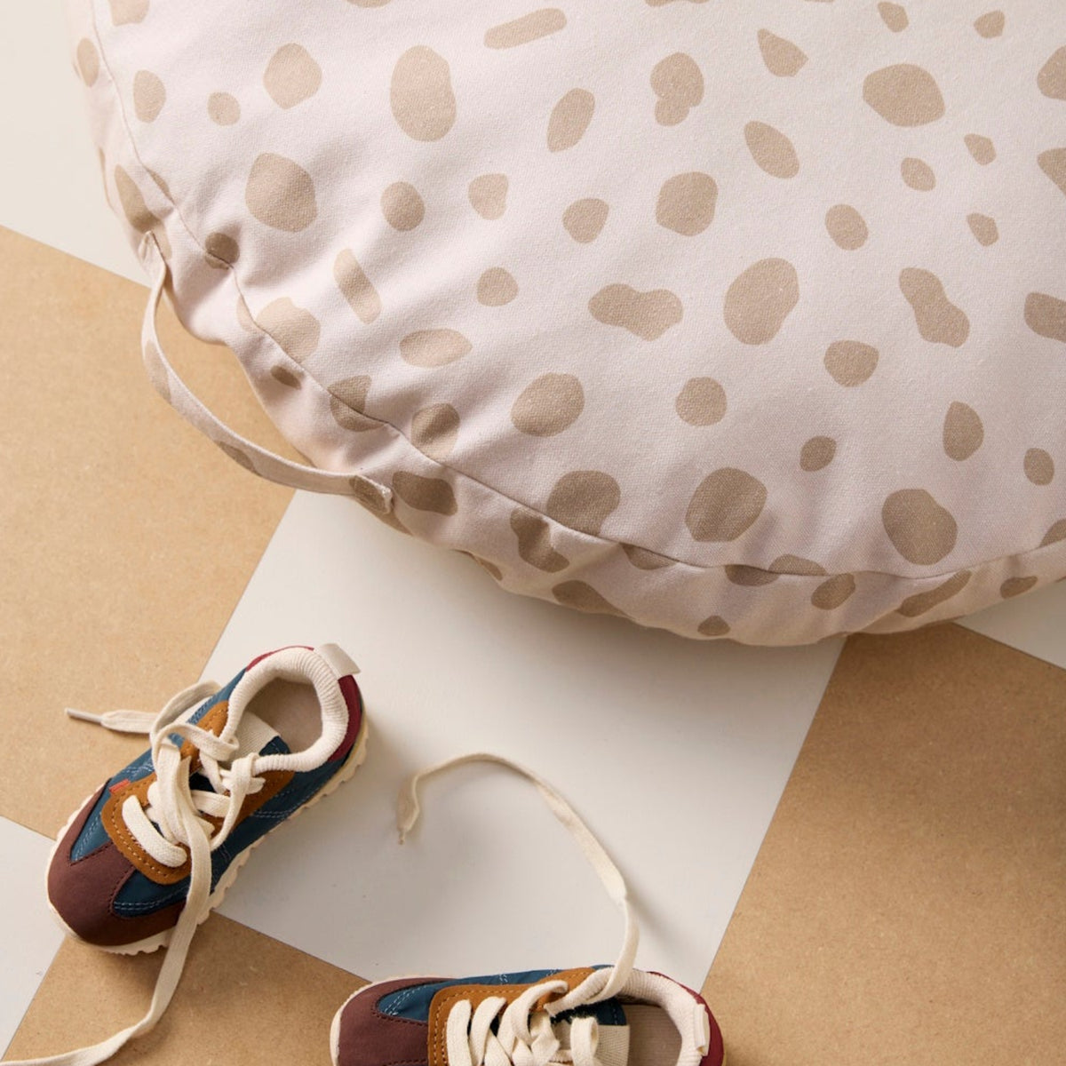 Round beige spotted floor cushion resting on a checkered floor next to a pair of kids' sneakers.