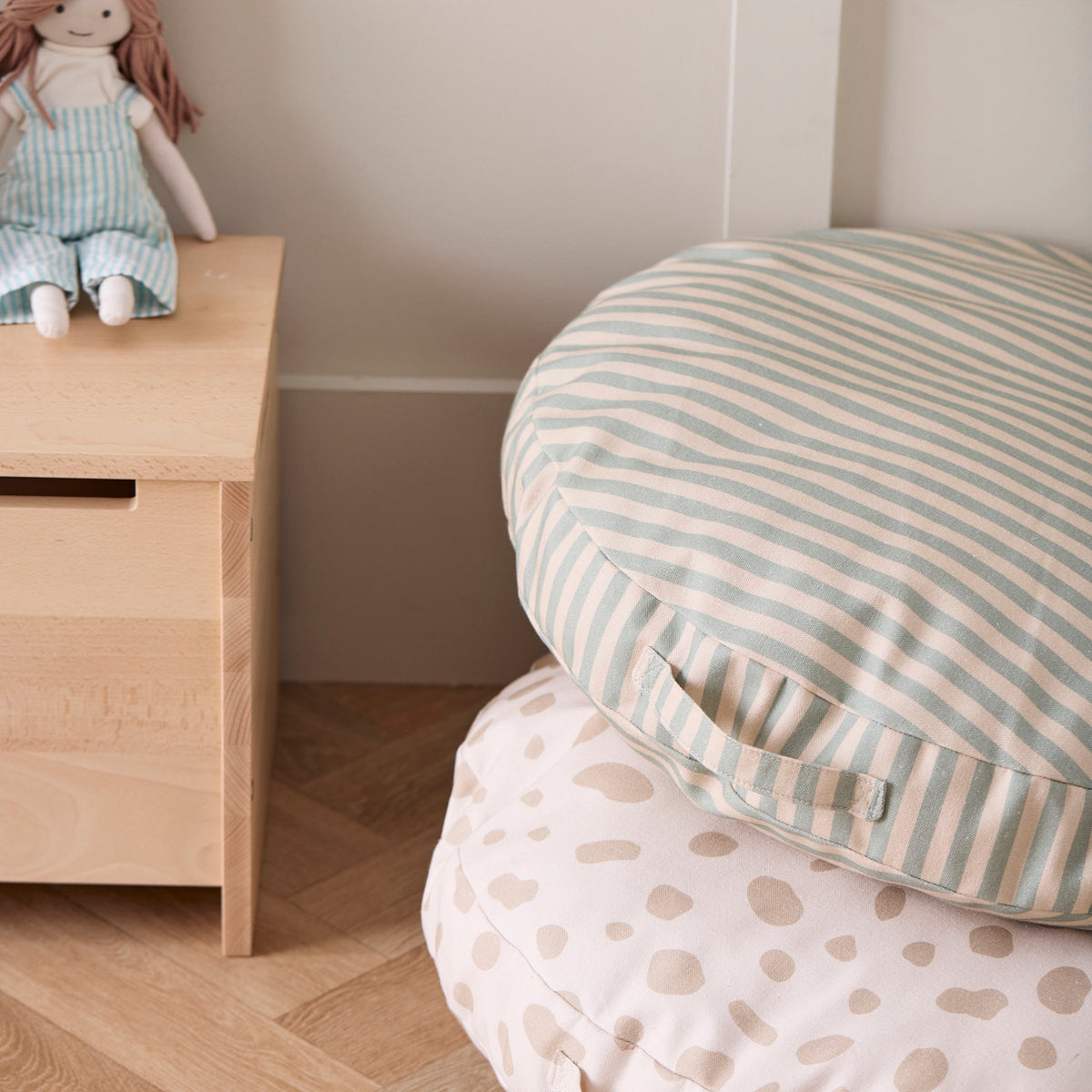 Green and cream striped floor cushion stacked on a spotted cushion next to a wooden chest with a rag doll.