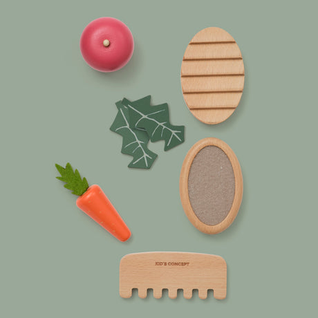 Wooden pet care toy set with grooming tools, a toy carrot, apple, and leaves on a green backdrop.
