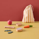 Wooden pet care toy set with play food and grooming tools on a table, with a striped bag behind.