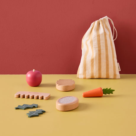 Wooden pet care toy set with play food and grooming tools on a table, with a striped bag behind.