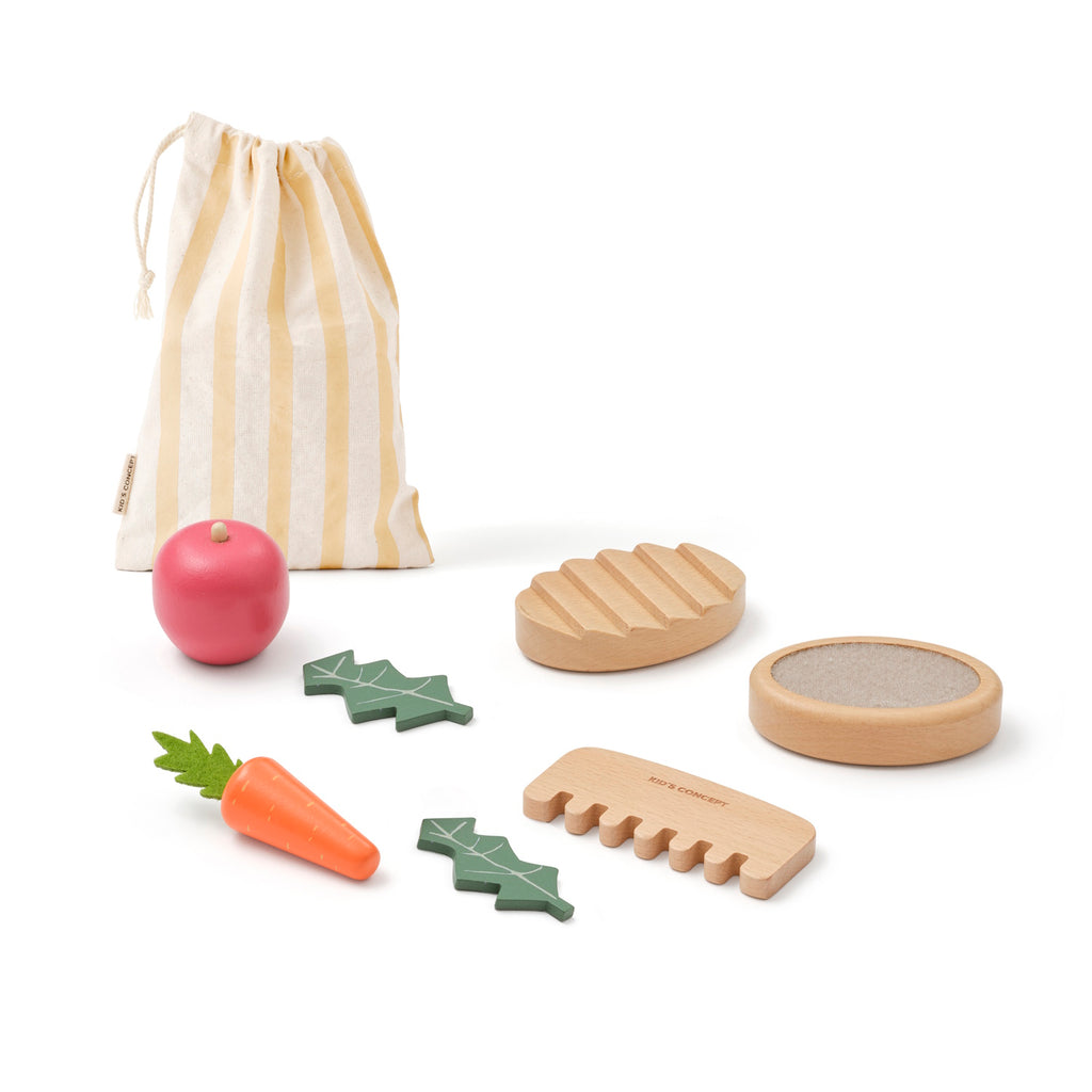 Toy kitchen set with wooden vegetables, bread, and accessories on a white background. Includes a striped pouch, red apple, carrot, and cutting board. Playful tone.